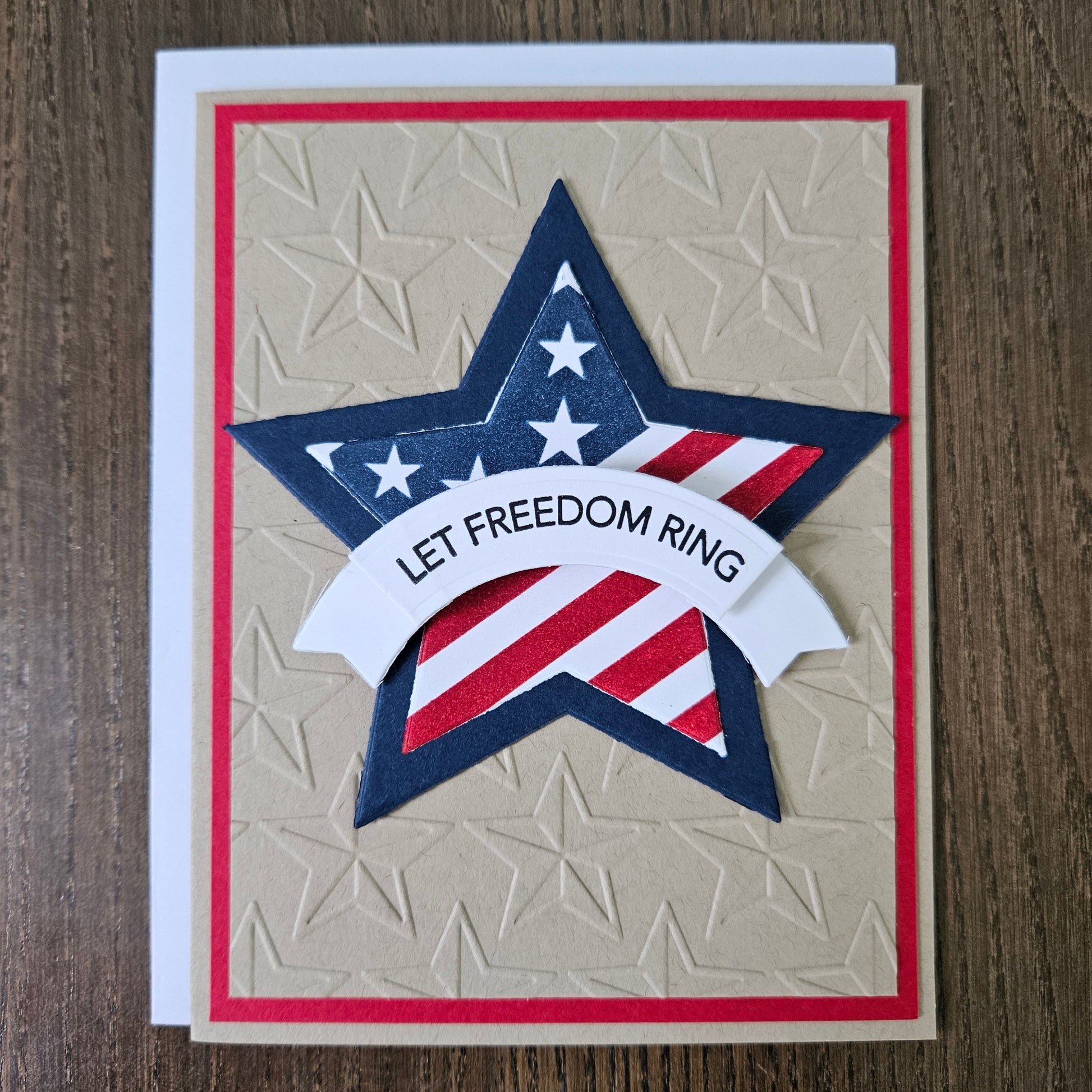 Handmade 4th of July card featuring a large layered star with an American flag design and a white dimensional banner reading Let Freedom Ring. The star is centered on a rustic kraft paper background deeply embossed with a repeating star pattern and framed by a thin red border layer.