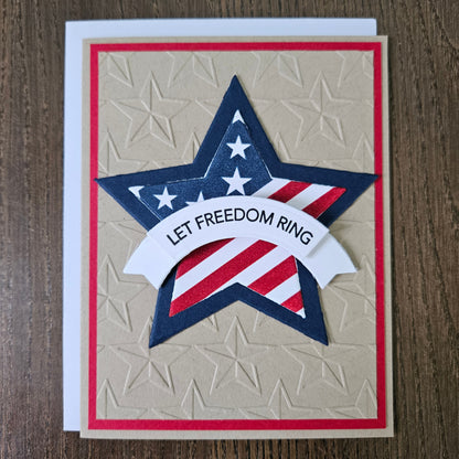 Handmade 4th of July card featuring a large layered star with an American flag design and a white dimensional banner reading Let Freedom Ring. The star is centered on a rustic kraft paper background deeply embossed with a repeating star pattern and framed by a thin red border layer.