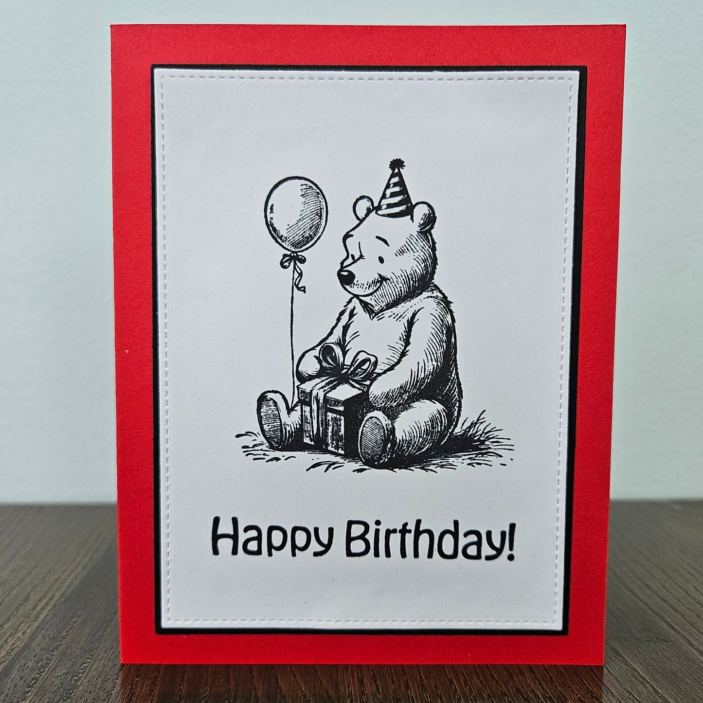 handmade A2 birthday card featuring a classic black ink storybook sketch illustration of Winnie the Pooh wearing a striped party hat while sitting with a wrapped gift box and a balloon, printed with the sentiment Happy Birthday!, finished with a stitched edge border, black mat, and red cardstock
