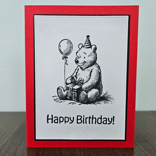 handmade A2 birthday card featuring a classic black ink storybook sketch illustration of Winnie the Pooh wearing a striped party hat while sitting with a wrapped gift box and a balloon, printed with the sentiment Happy Birthday!, finished with a stitched edge border, black mat, and red cardstock
