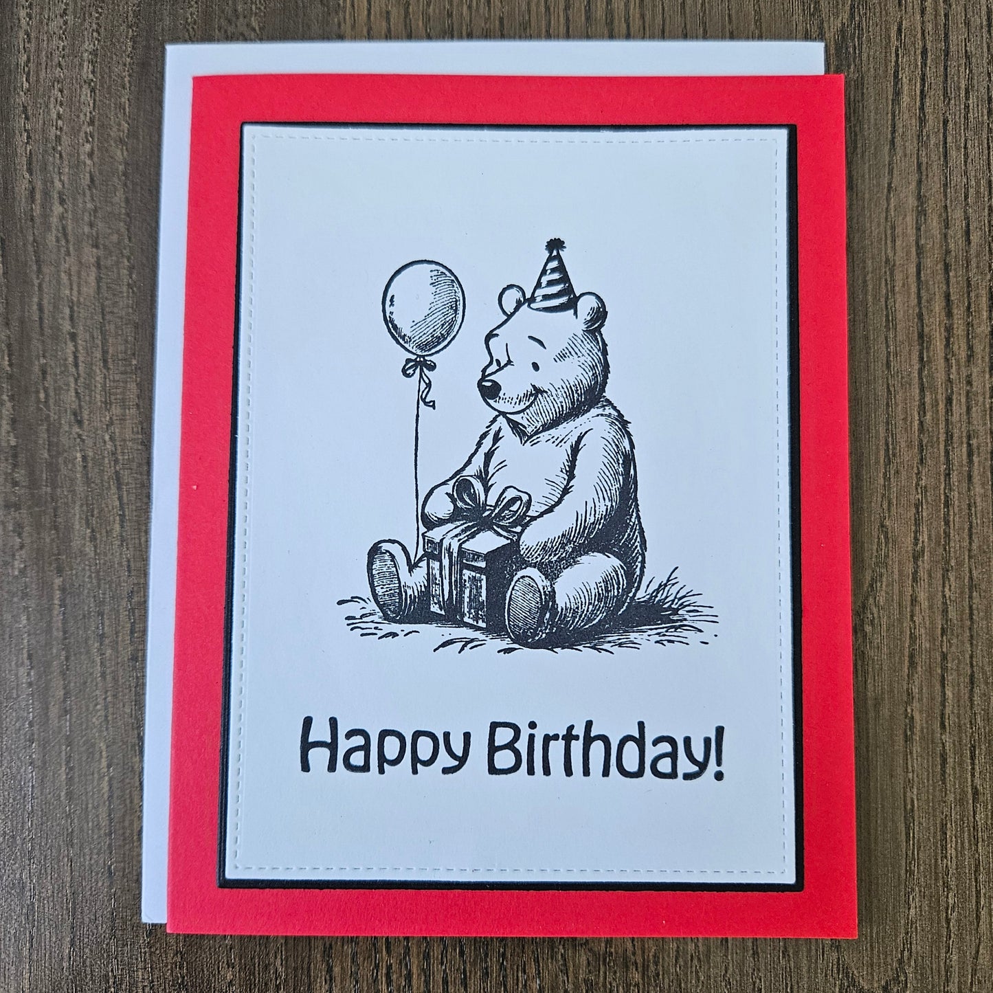 handmade A2 birthday card featuring a classic black ink storybook sketch illustration of Winnie the Pooh wearing a striped party hat while sitting with a wrapped gift box and a balloon, printed with the sentiment Happy Birthday!, finished with a stitched edge border, black mat, and red cardstock