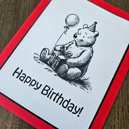 handmade A2 birthday card featuring a classic black ink storybook sketch illustration of Winnie the Pooh wearing a striped party hat while sitting with a wrapped gift box and a balloon, printed with the sentiment Happy Birthday!, finished with a stitched edge border, black mat, and red cardstock