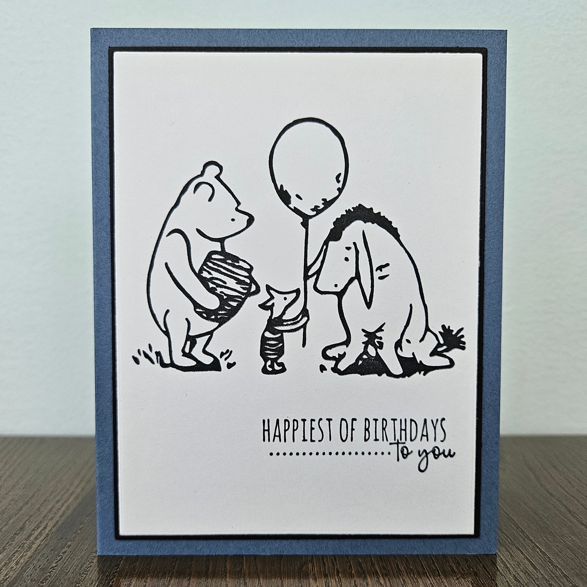 a handmade A2 birthday card featuring a classic black ink storybook sketch illustration of Winnie the Pooh with a honey pot, Piglet holding a balloon, and Eeyore standing together, printed with the sentiment Happiest of Birthdays To you, finished with a dark mat and blue cardstock frame