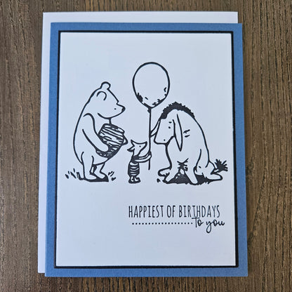 a handmade A2 birthday card featuring a classic black ink storybook sketch illustration of Winnie the Pooh with a honey pot, Piglet holding a balloon, and Eeyore standing together, printed with the sentiment Happiest of Birthdays To you, finished with a dark mat and blue cardstock frame