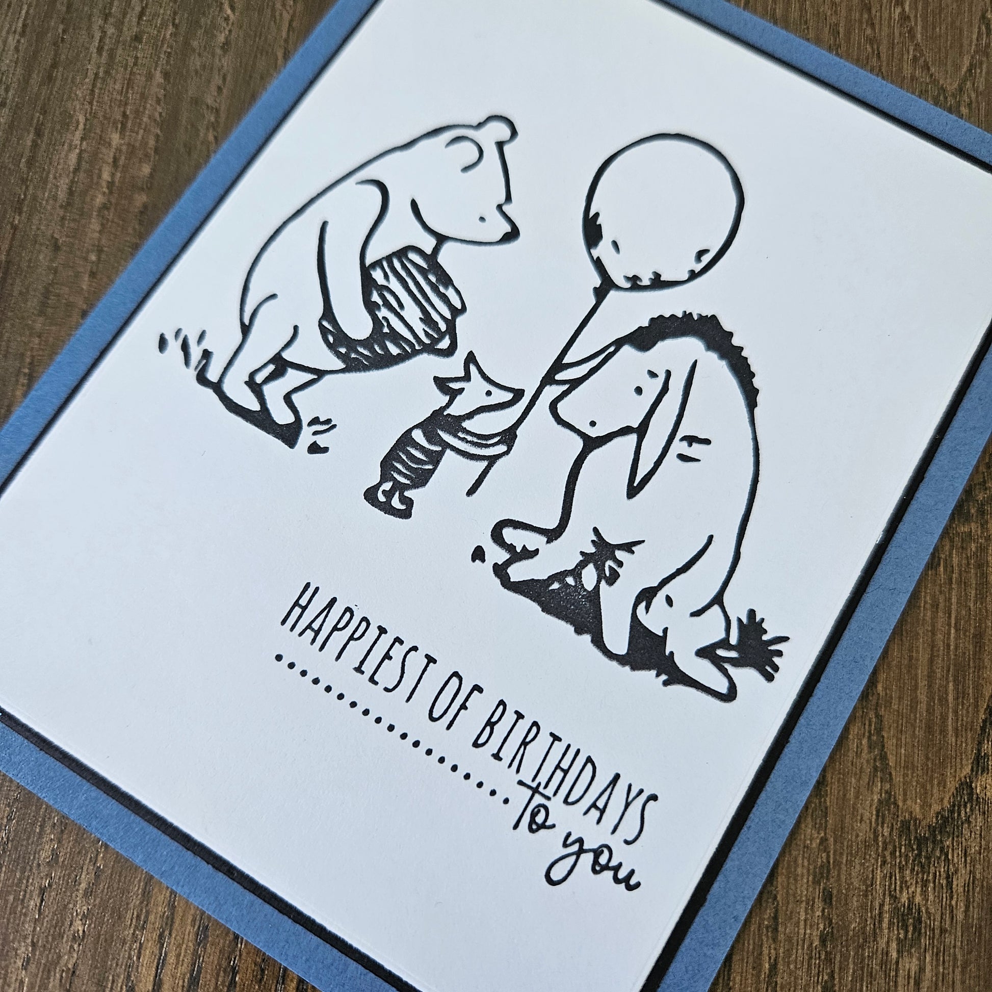 a handmade A2 birthday card featuring a classic black ink storybook sketch illustration of Winnie the Pooh with a honey pot, Piglet holding a balloon, and Eeyore standing together, printed with the sentiment Happiest of Birthdays To you, finished with a dark mat and blue cardstock frame