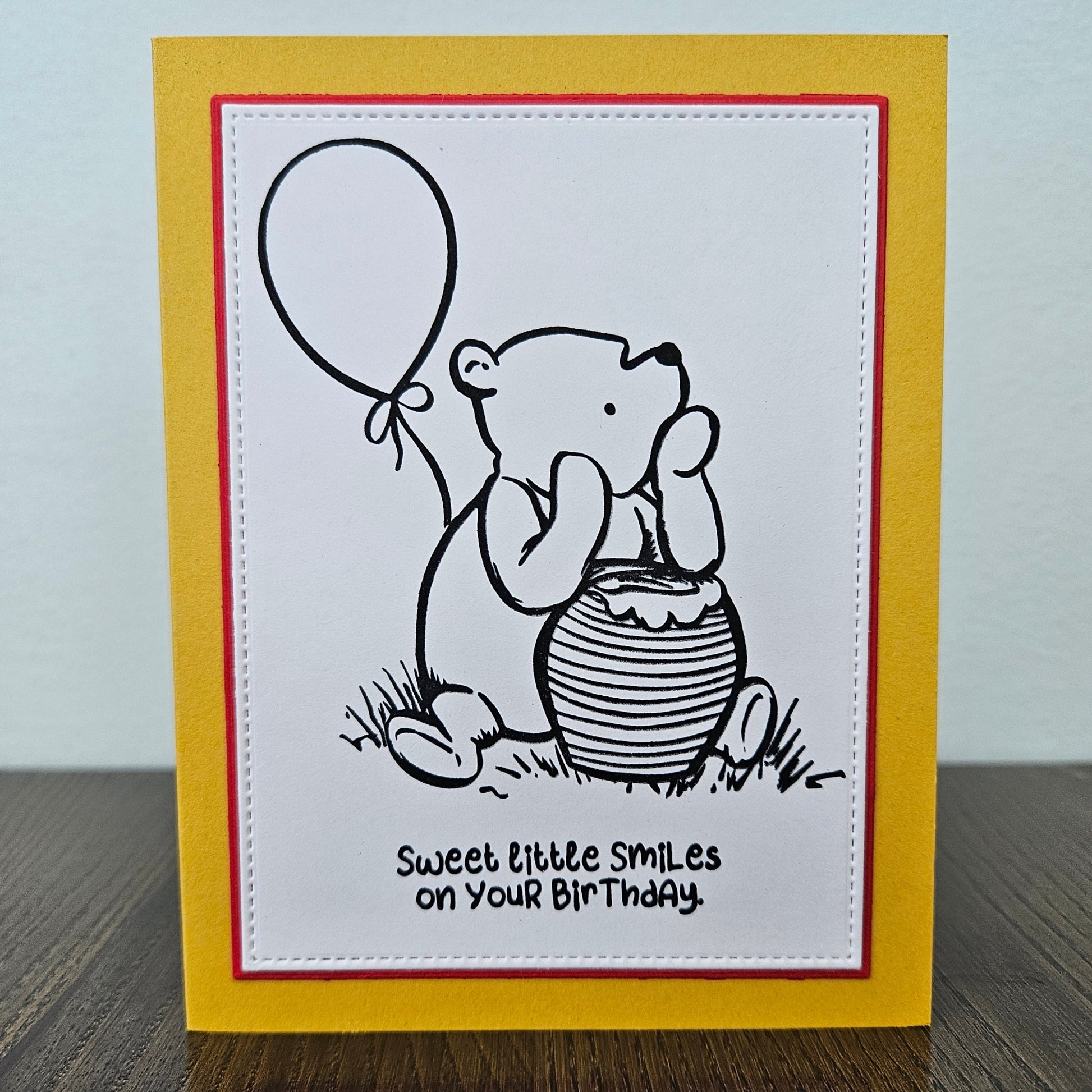 a handmade A2 birthday card featuring a classic black ink storybook sketch illustration of Winnie the Pooh sitting on the grass next to a large honey pot with a balloon floating behind him, printed with the sentiment Sweet little smiles on your birthday, finished with a stitched edge border, red mat, and bright yellow cardstock frame