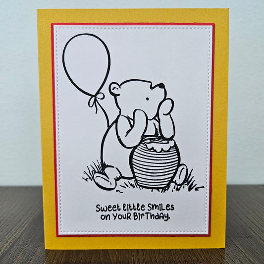 a handmade A2 birthday card featuring a classic black ink storybook sketch illustration of Winnie the Pooh sitting on the grass next to a large honey pot with a balloon floating behind him, printed with the sentiment Sweet little smiles on your birthday, finished with a stitched edge border, red mat, and bright yellow cardstock frame