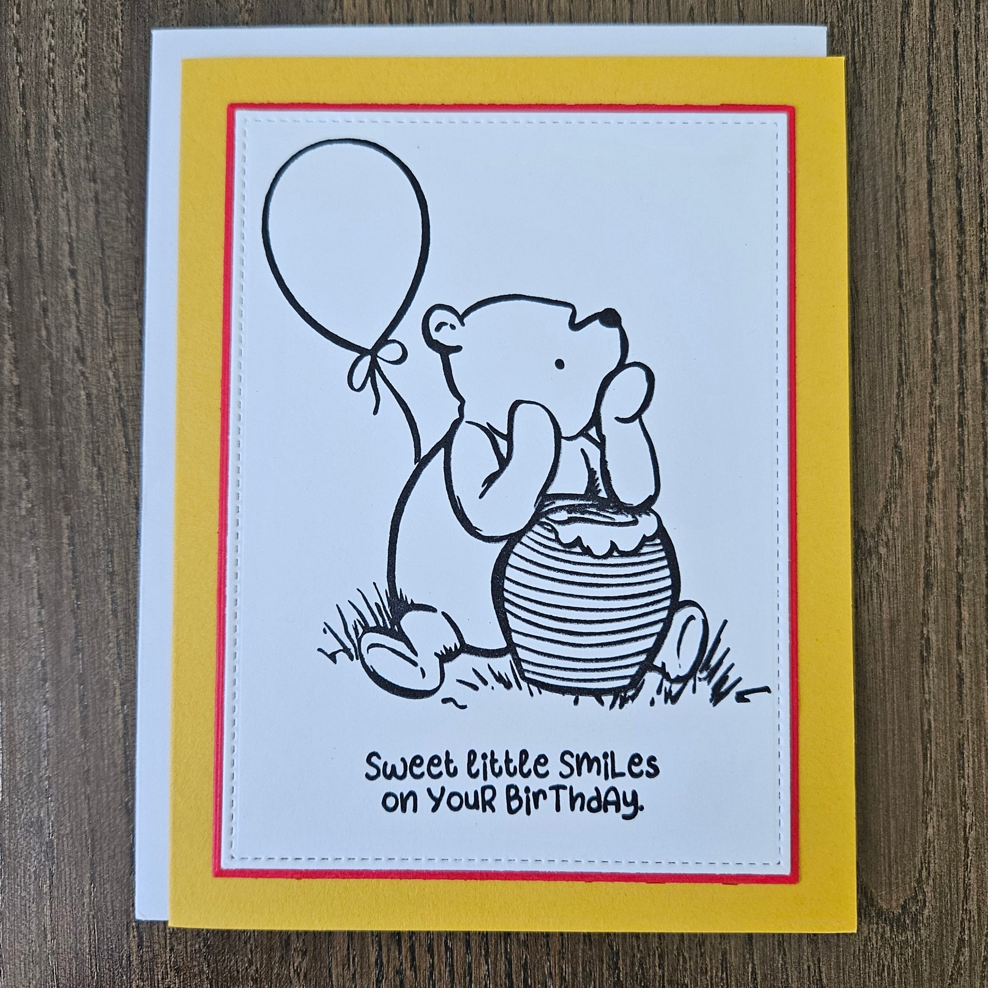 a handmade A2 birthday card featuring a classic black ink storybook sketch illustration of Winnie the Pooh sitting on the grass next to a large honey pot with a balloon floating behind him, printed with the sentiment Sweet little smiles on your birthday, finished with a stitched edge border, red mat, and bright yellow cardstock frame