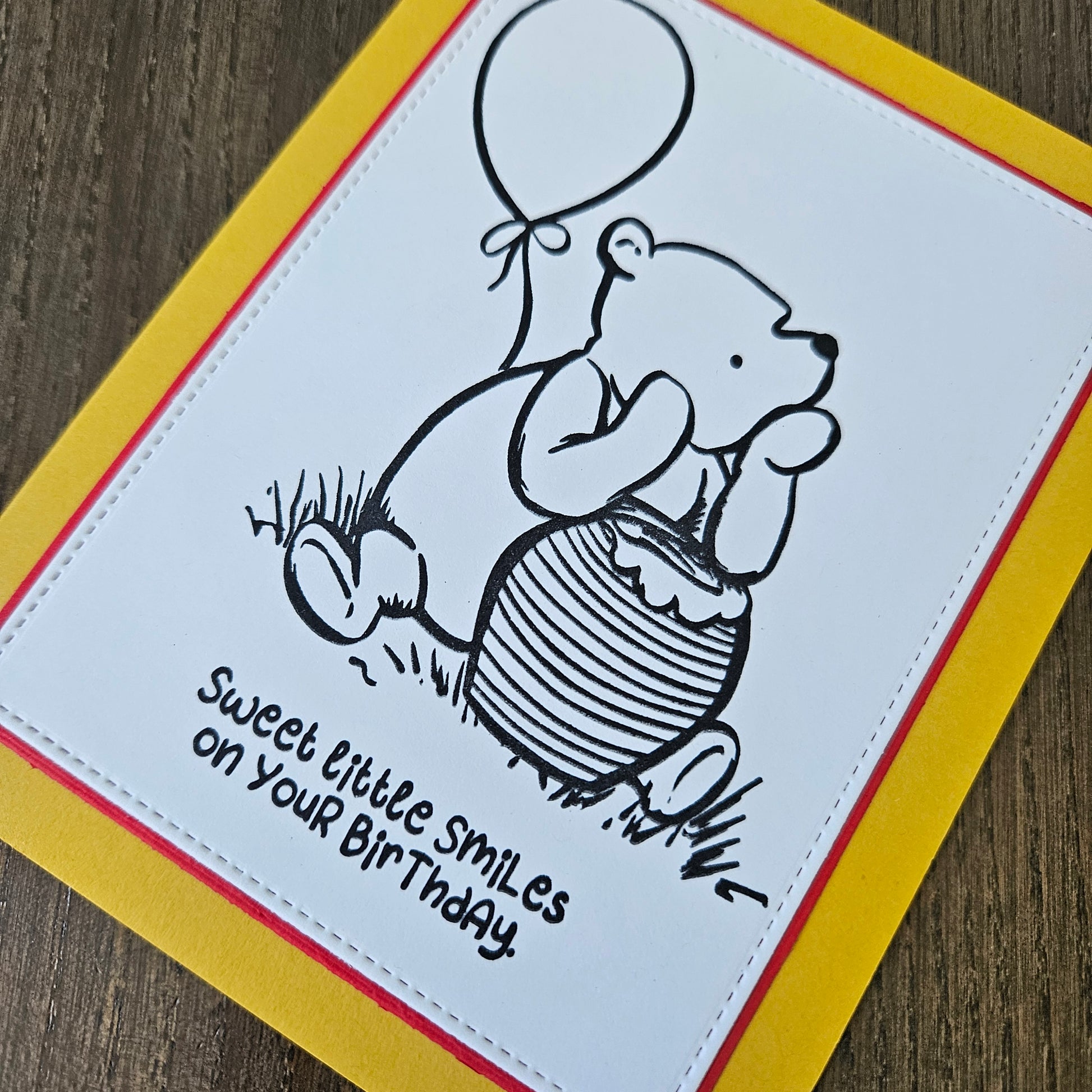 a handmade A2 birthday card featuring a classic black ink storybook sketch illustration of Winnie the Pooh sitting on the grass next to a large honey pot with a balloon floating behind him, printed with the sentiment Sweet little smiles on your birthday, finished with a stitched edge border, red mat, and bright yellow cardstock frame