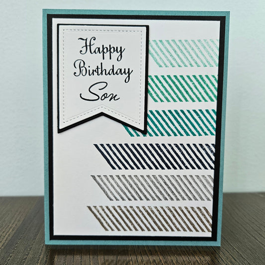 a handmade A2 card featuring a modern geometric design with multi-colored diagonal striped blocks and a stitched white pennant banner that reads Happy Birthday Son, finished with a crisp black mat and teal cardstock