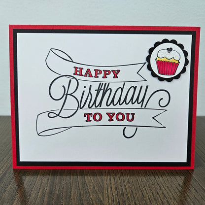 Front of a handmade A2 birthday card featuring a white background panel with a large sketched ribbon banner reading Happy Birthday To You accented in red ink, accented with a hand-colored cupcake with red frosting in a scalloped black circle mat, finished with a black inner border and bright red cardstock frame