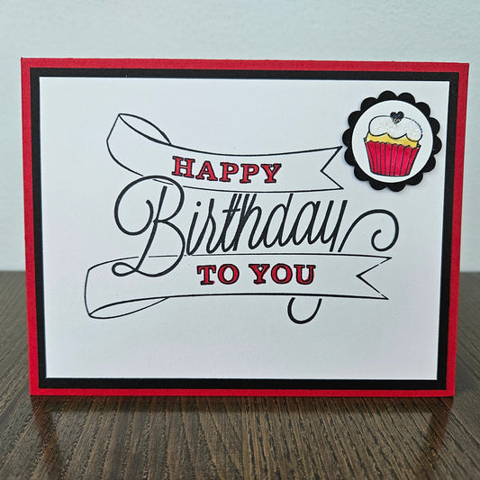 Front of a handmade A2 birthday card featuring a white background panel with a large sketched ribbon banner reading Happy Birthday To You accented in red ink, accented with a hand-colored cupcake with red frosting in a scalloped black circle mat, finished with a black inner border and bright red cardstock frame