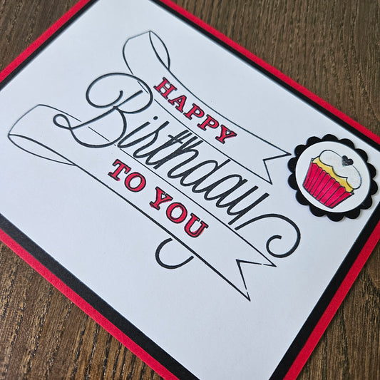 A square handmade-style birthday card with a white center, black and red border, and a cupcake illustration near the top. The card reads “Happy Birthday to You” in black and red lettering, photographed on a brown wood surface.