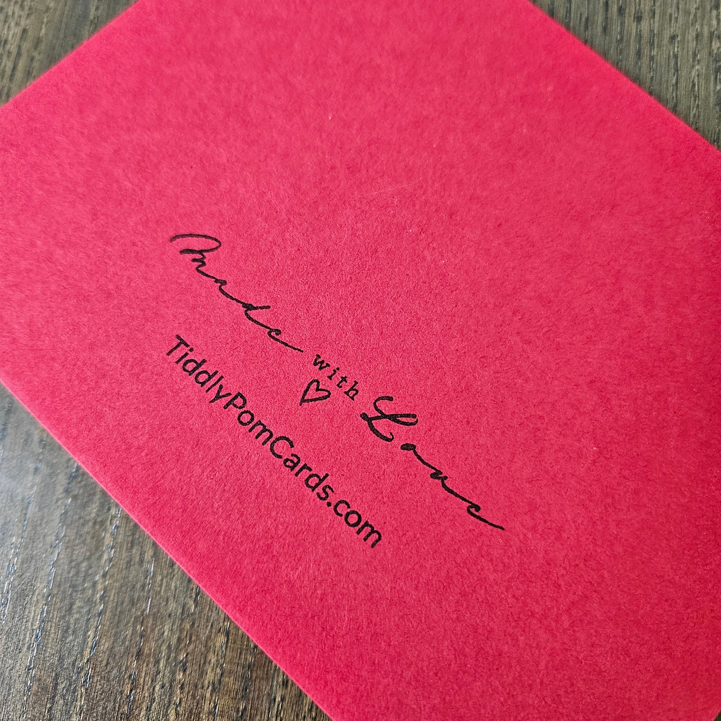 A close-up of a textured pinkish-red card or envelope corner with black printed script reading 'Made with Love' and 'TiddlyPomCards.com', placed on a wood surface.