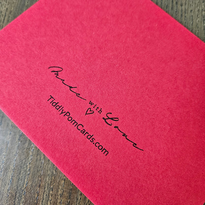 A close-up of a textured pinkish-red card or envelope corner with black printed script reading 'Made with Love' and 'TiddlyPomCards.com', placed on a wood surface.