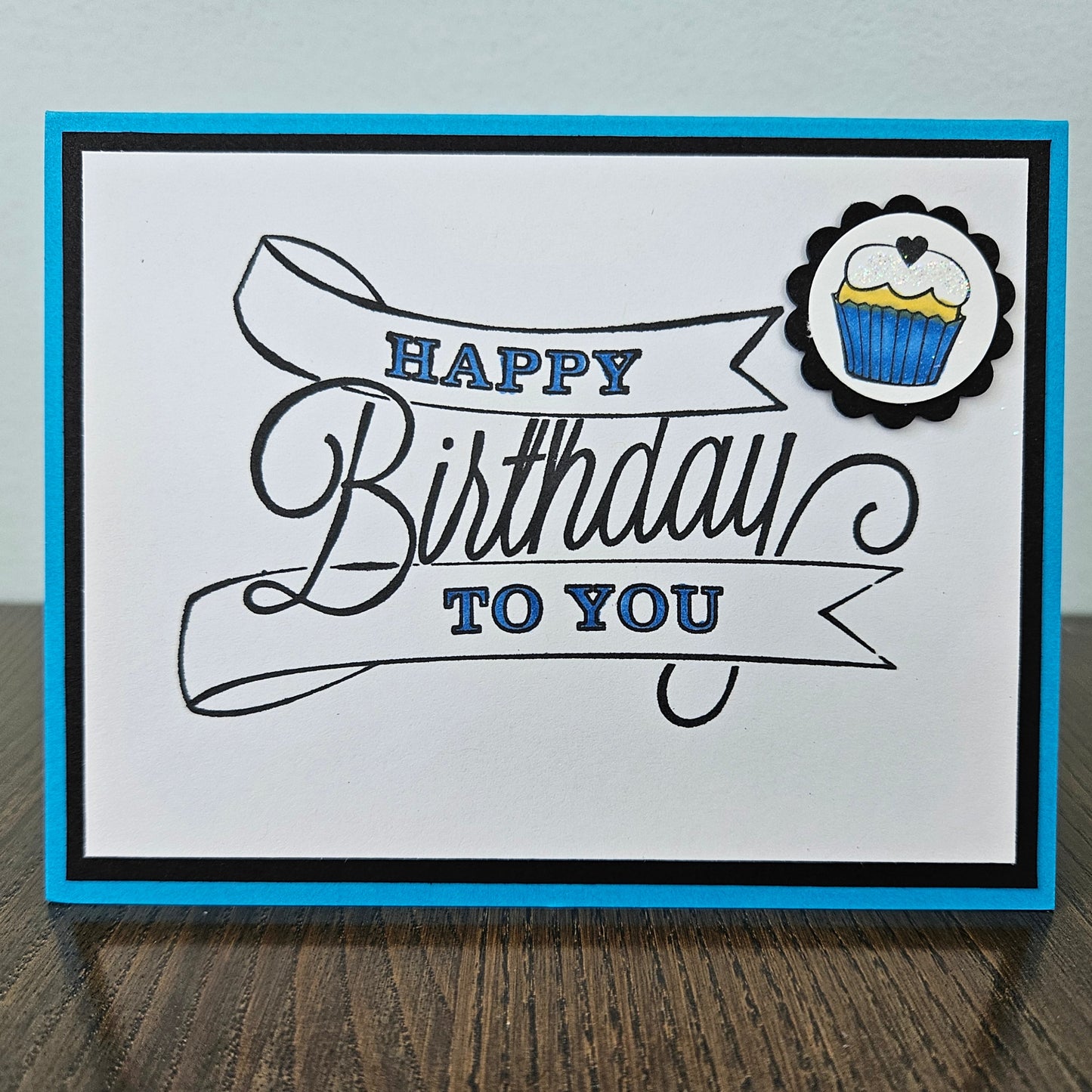 A handmade square greeting card with a white center panel, black and blue border, and the words “HAPPY Birthday TO YOU” in black and blue script. A small cupcake sticker is in the top right corner, and the card is photographed standing on a wooden surface against a light wall.
