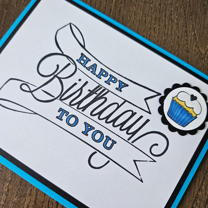 A square birthday card with a white face, black script lettering that reads 'Happy Birthday to You,' a blue border, and a small cupcake illustration near the top, photographed on a wooden surface.