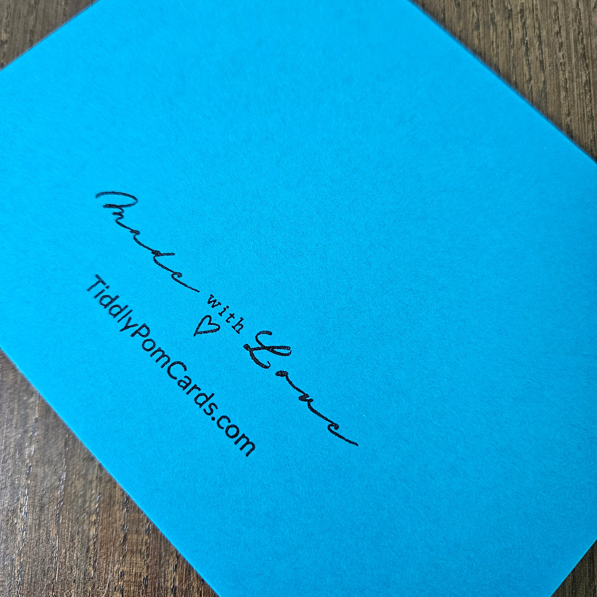 A bright blue greeting card or card back with black printed script reading 'Proud with Love' and 'TiddlyPomCards.com', photographed on a wooden surface.