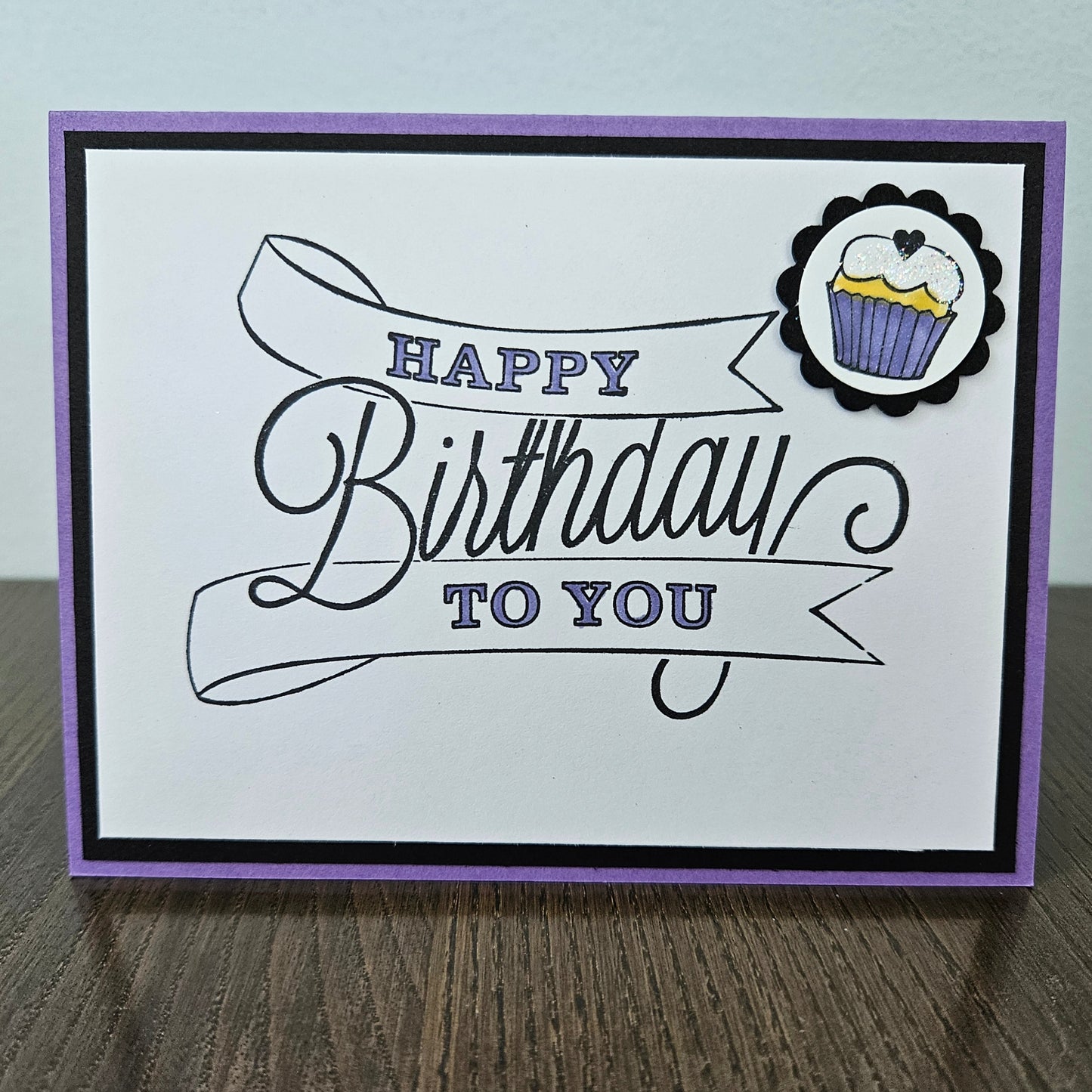 A handmade square greeting card with a purple border, black and purple 'Happy Birthday to You' script text, and a small cupcake sticker in the top right corner, displayed on a wooden surface.
