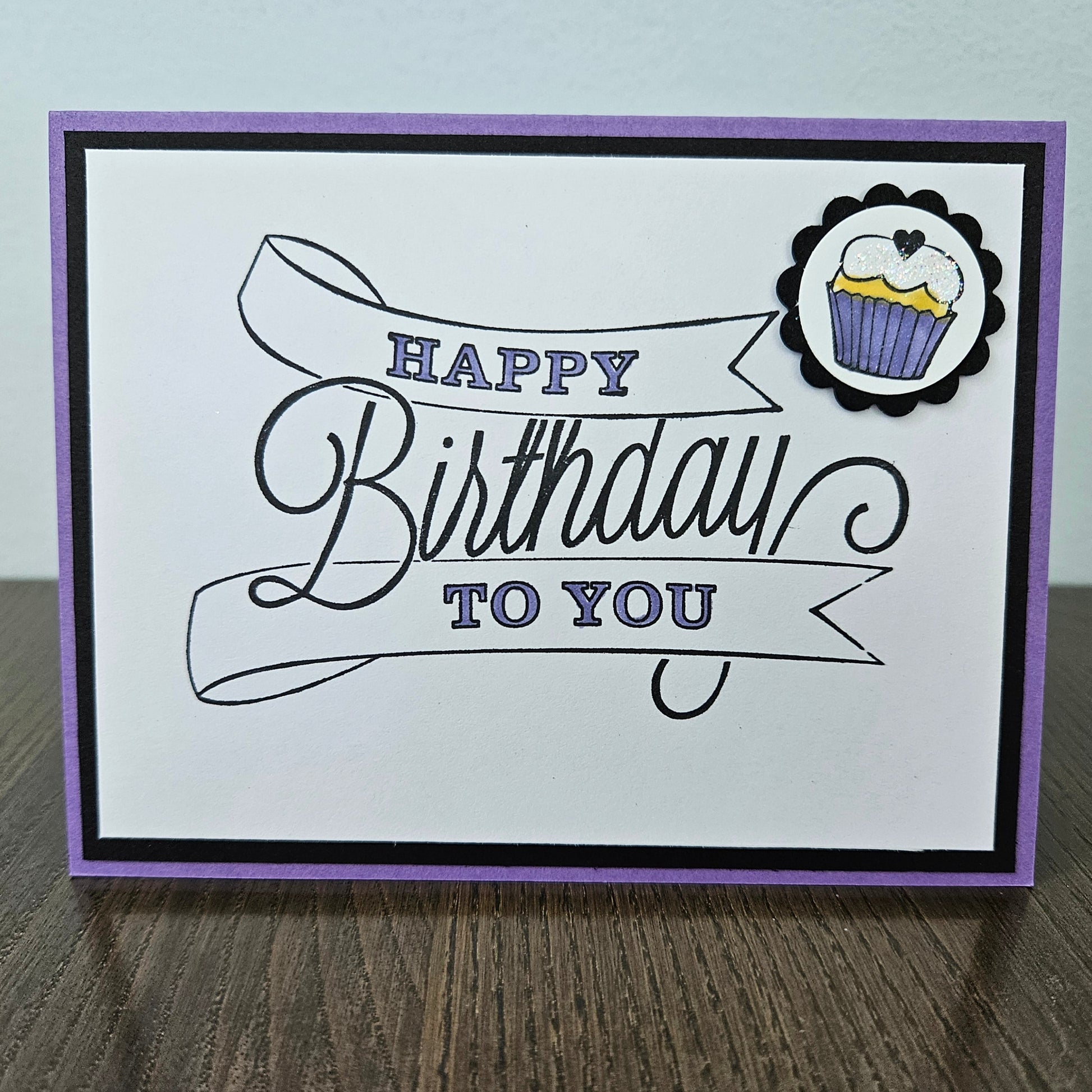 A handmade square greeting card with a purple border, black and purple 'Happy Birthday to You' script text, and a small cupcake sticker in the top right corner, displayed on a wooden surface.