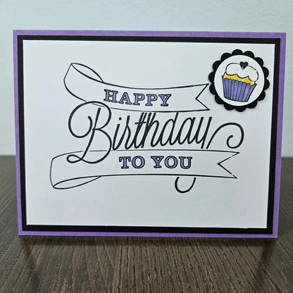 A handmade square greeting card with a purple border, black and purple 'Happy Birthday to You' script text, and a small cupcake sticker in the top right corner, displayed on a wooden surface.