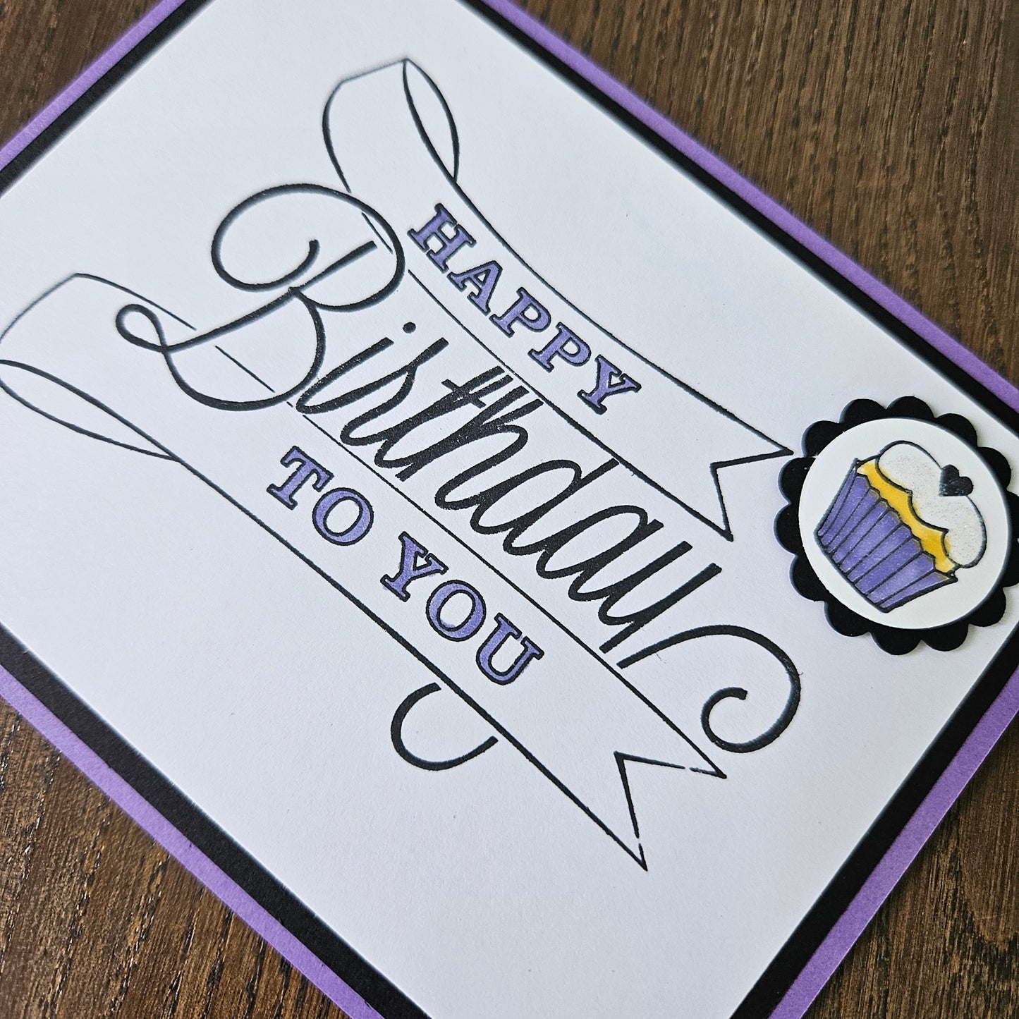 A square handmade greeting card with a white face, black and purple border, and a decorative cupcake illustration at the top. The card reads 'Happy Birthday to You' in black and purple script, photographed on a wooden surface.