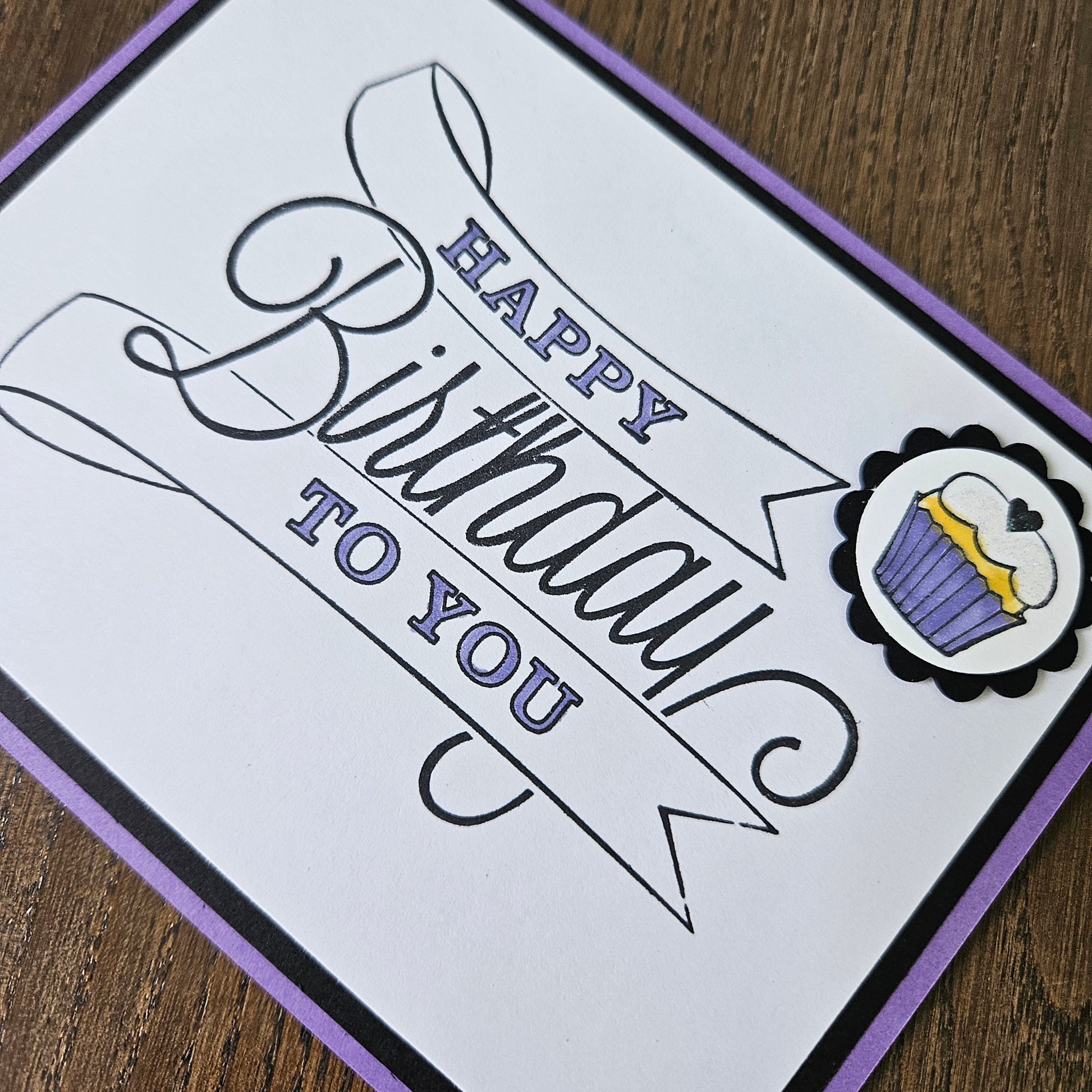 A square handmade greeting card with a white face, black and purple border, and a decorative cupcake illustration at the top. The card reads 'Happy Birthday to You' in black and purple script, photographed on a wooden surface.