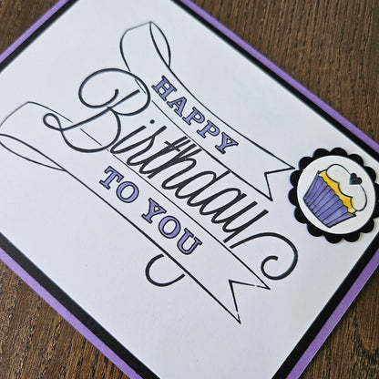 A square handmade greeting card with a white face, black and purple border, and a decorative cupcake illustration at the top. The card reads 'Happy Birthday to You' in black and purple script, photographed on a wooden surface.