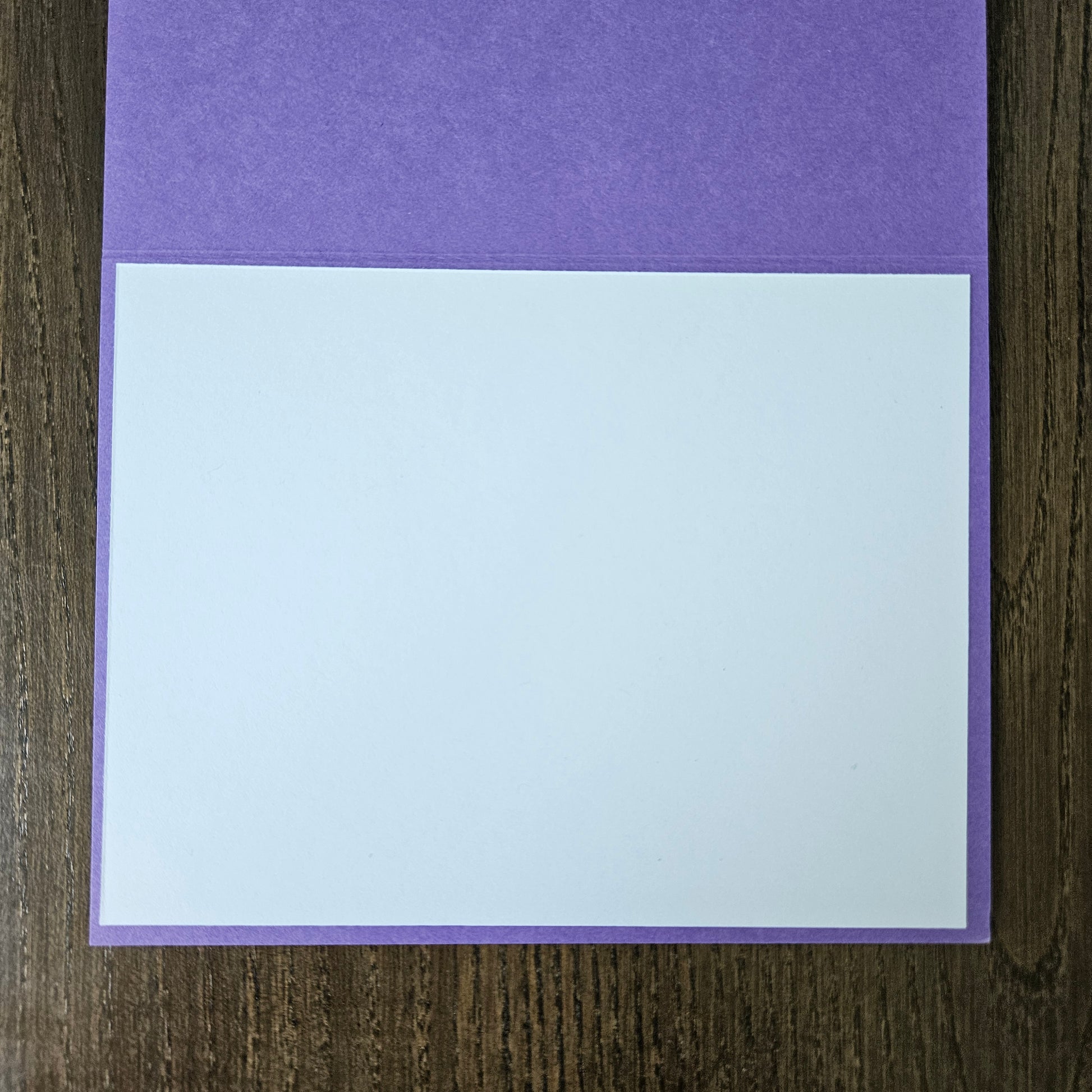 A blank white rectangular card or paper sheet placed on top of a purple textured cover or folder, photographed from above on a dark wood surface.