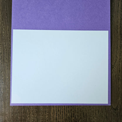 A blank white rectangular card or paper sheet placed on top of a purple textured cover or folder, photographed from above on a dark wood surface.