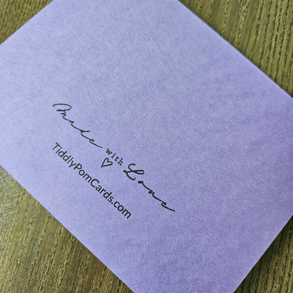 A square lavender purple card with black printed text reading 'Made with Love' and 'TidilyPomCards.com', photographed on a wood surface.