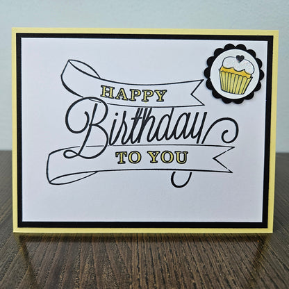 A square handmade greeting card with 'HAPPY Birthday TO YOU' in black and gold lettering, featuring a small cupcake sticker in the top right corner, displayed on a dark wood surface against a light gray wall.