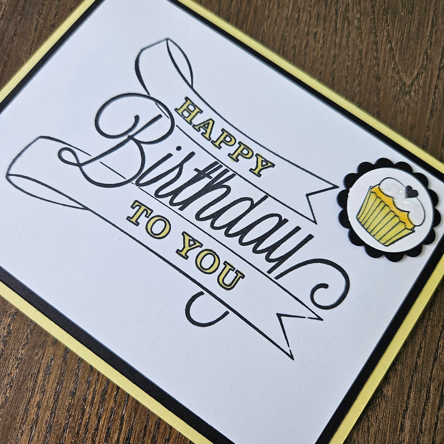 A close-up of a square birthday card with a white center, black and gold decorative script reading 'Happy Birthday to You,' a cupcake illustration at the top, and a gold border, placed on a brown wood surface.