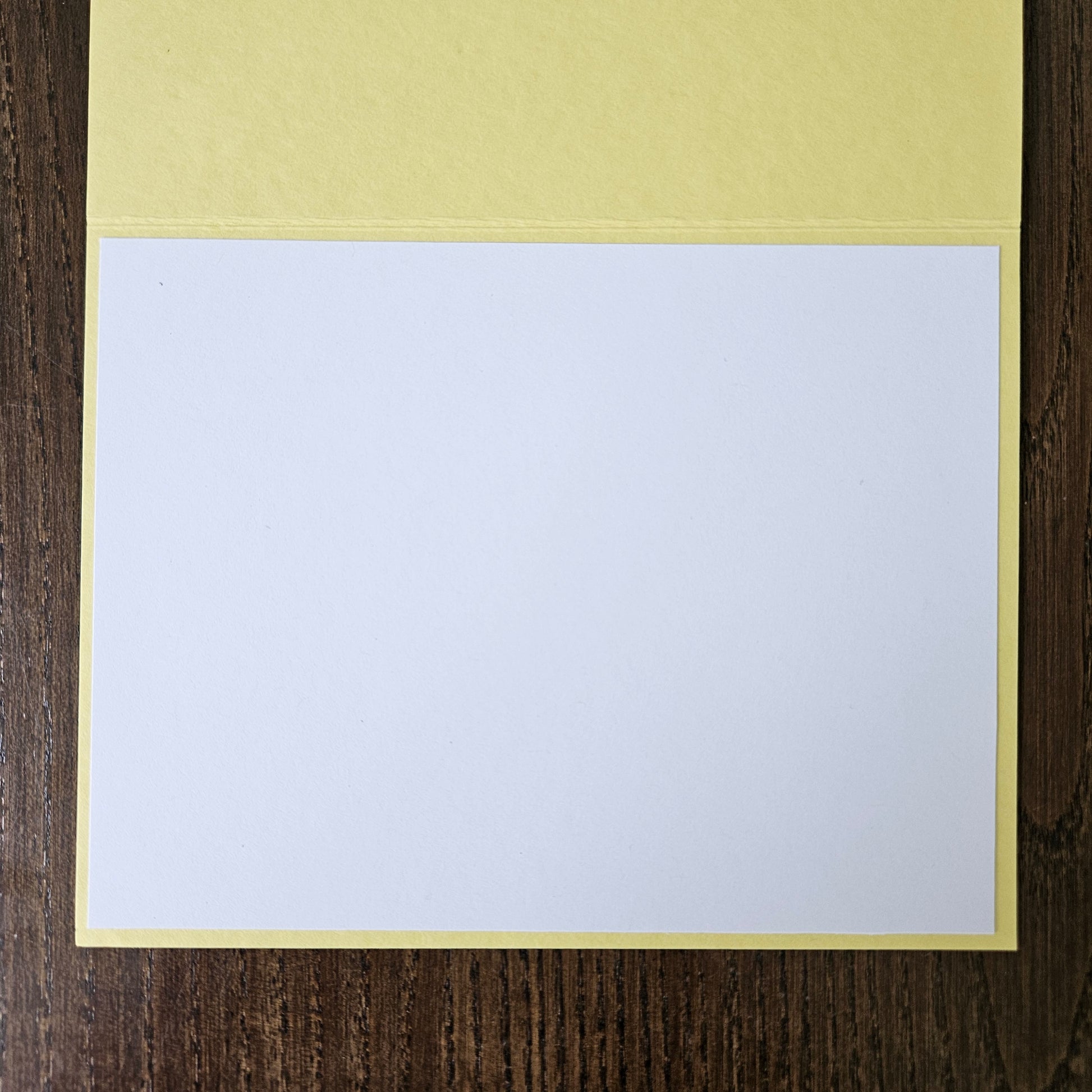 A blank white card or paper sheet with a pale yellow border, placed on a dark brown wood surface.