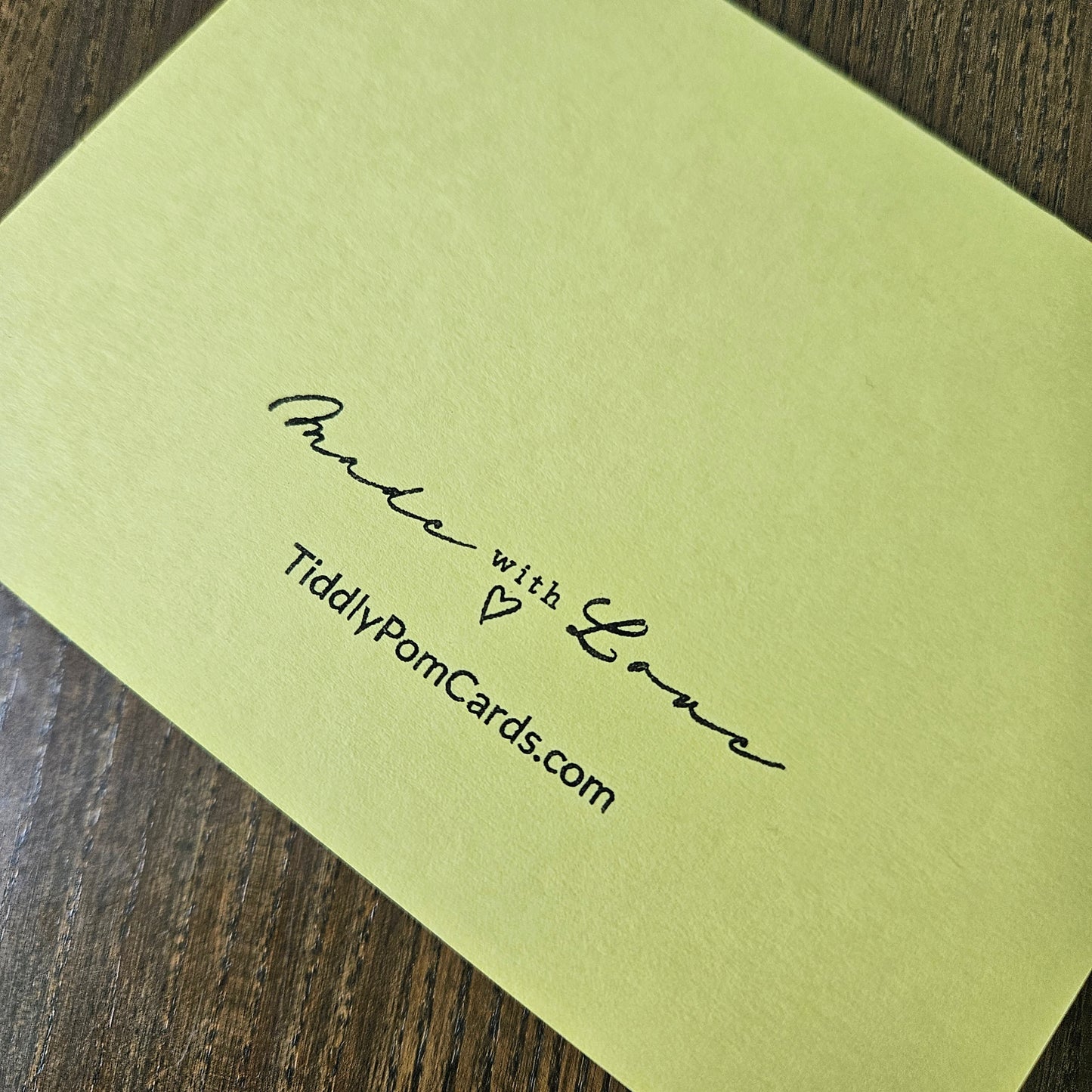 A pale yellow-green greeting card with black printed text reading 'Made with Love' and 'TiddyPomCards.com', photographed on a dark wood surface.