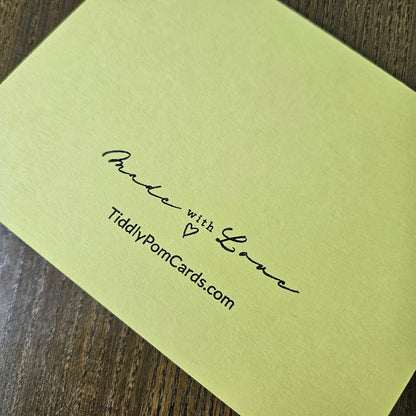 A pale yellow-green greeting card with black printed text reading 'Made with Love' and 'TiddyPomCards.com', photographed on a dark wood surface.