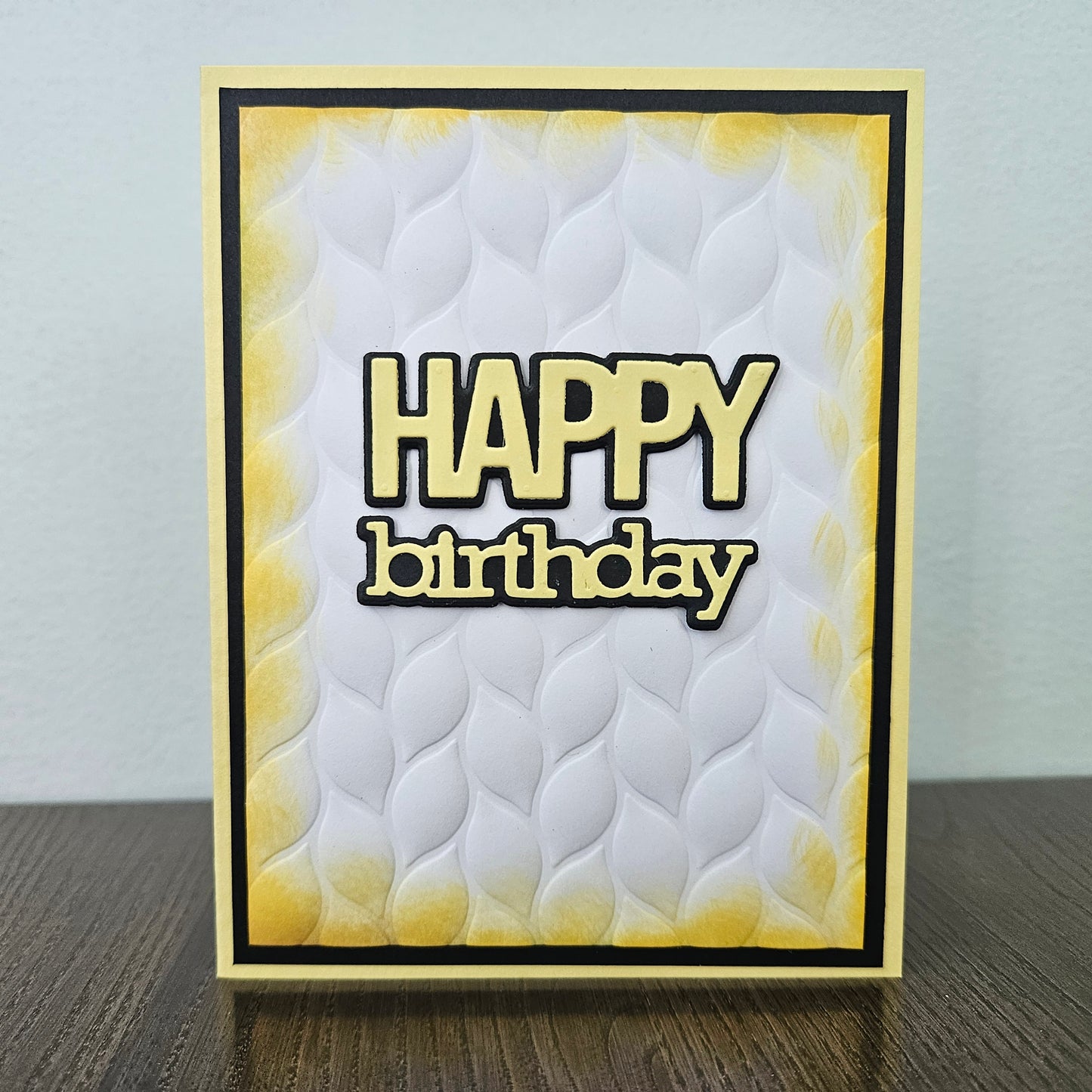 Front of a handmade 5x7 birthday card featuring an embossed white leaf-pattern background panel with hand-inked yellow edges, featuring bold die-cut text that reads HAPPY birthday in yellow with a crisp black outline, finished with a black inner mat and light yellow cardstock frame