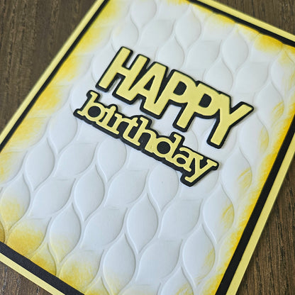 A close-up of a handmade greeting card featuring the words 'HAPPY birthday' in raised yellow letters with black outlines, set against a white embossed wavy background with yellow and black border details, resting on a wooden surface.