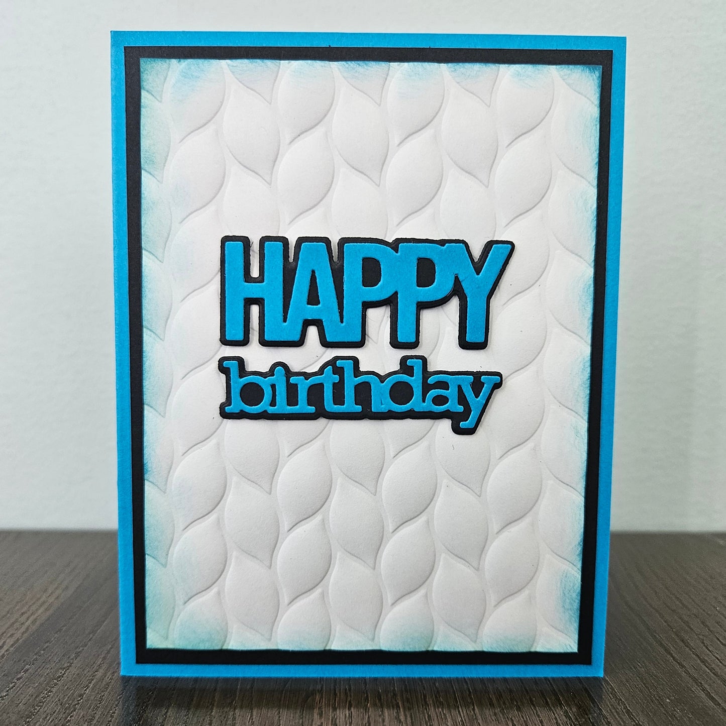 Front of the handmade 5x7 birthday card featuring an embossed white leaf-pattern background panel with hand-inked blue edges, featuring bold die-cut text that reads HAPPY birthday in blue with a crisp black outline, finished with a black inner mat and vibrant blue cardstock frame
