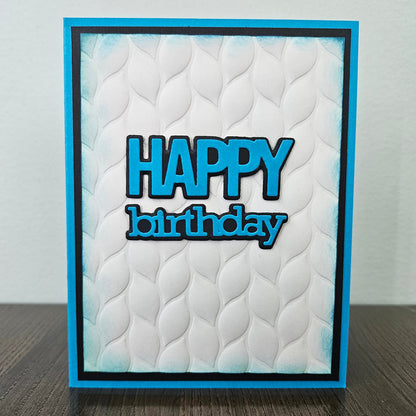 Front of the handmade 5x7 birthday card featuring an embossed white leaf-pattern background panel with hand-inked blue edges, featuring bold die-cut text that reads HAPPY birthday in blue with a crisp black outline, finished with a black inner mat and vibrant blue cardstock frame
