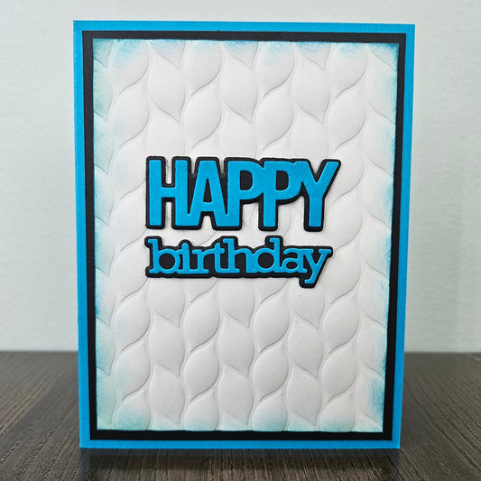 Front of the handmade 5x7 birthday card featuring an embossed white leaf-pattern background panel with hand-inked blue edges, featuring bold die-cut text that reads HAPPY birthday in blue with a crisp black outline, finished with a black inner mat and vibrant blue cardstock frame