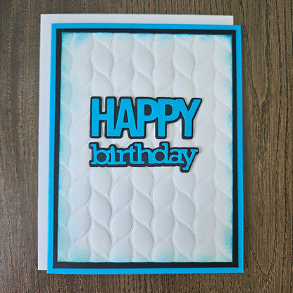 Front of the handmade 5x7 birthday card featuring an embossed white leaf-pattern background panel with hand-inked blue edges, featuring bold die-cut text that reads HAPPY birthday in blue with a crisp black outline, finished with a black inner mat and vibrant blue cardstock frame