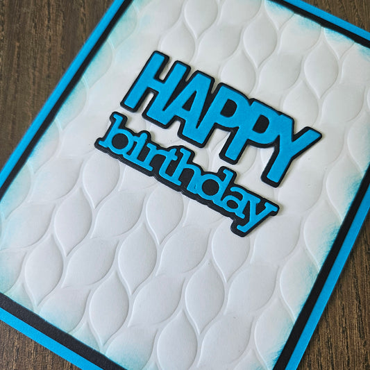A close-up of a handmade greeting card featuring a raised white wave pattern, a bright blue border, and a die-cut 'HAPPY birthday' text element in blue with a black outline, resting on a dark wood surface.