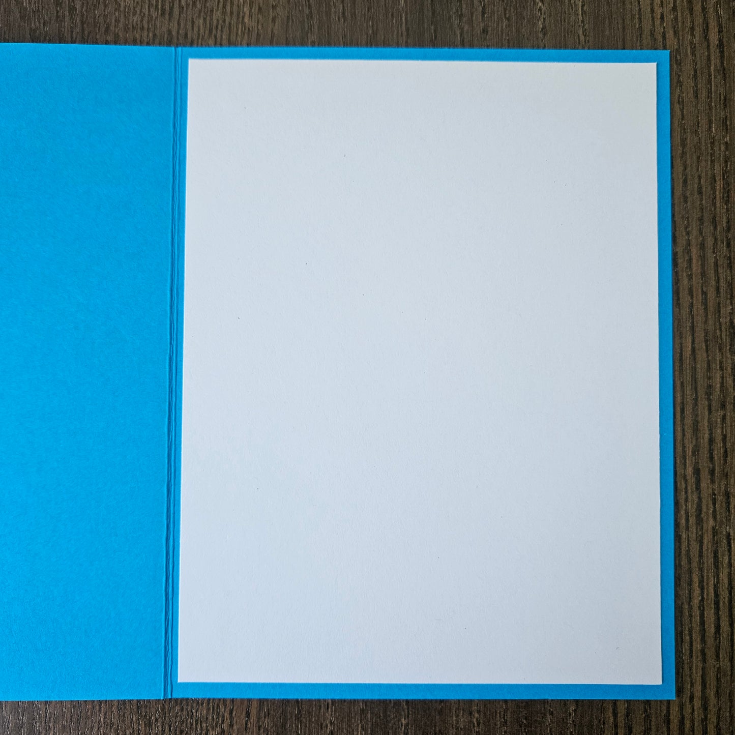 A square blue greeting card or paper folder with a blank white insert, photographed from above on a dark wood surface.