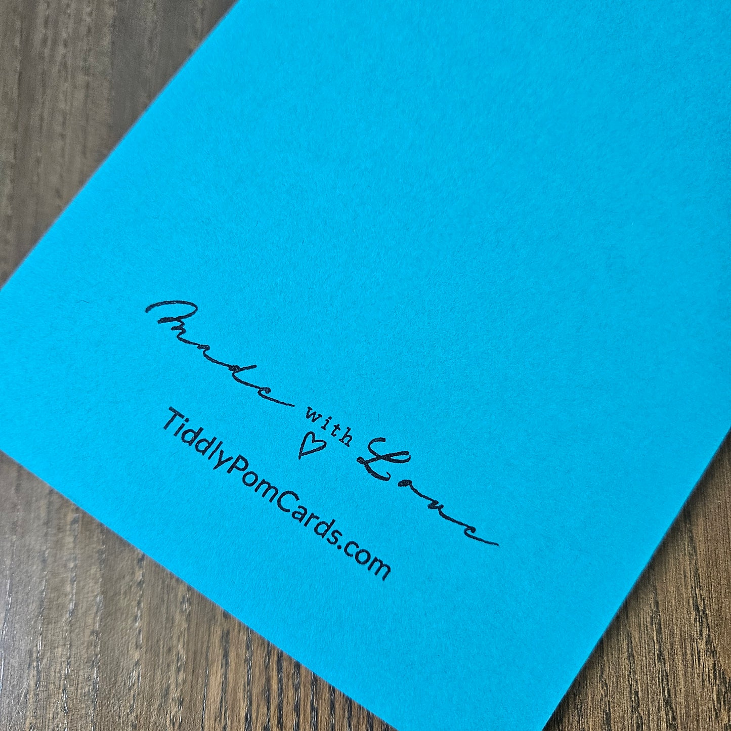 A close-up of a bright blue personalized pom card on a wooden surface, printed with the text 'Made with Love' and 'TiddlyPomCards.com'.