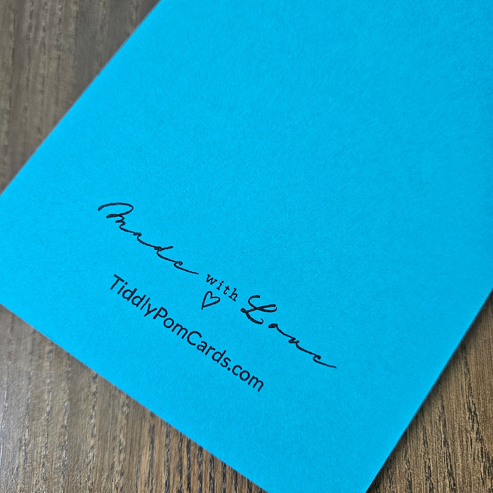 A close-up of a bright blue personalized pom card on a wooden surface, printed with the text 'Made with Love' and 'TiddlyPomCards.com'.