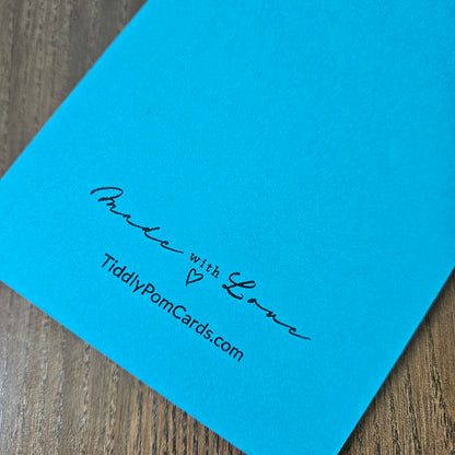 A close-up of a bright blue personalized pom card on a wooden surface, printed with the text 'Made with Love' and 'TiddlyPomCards.com'.