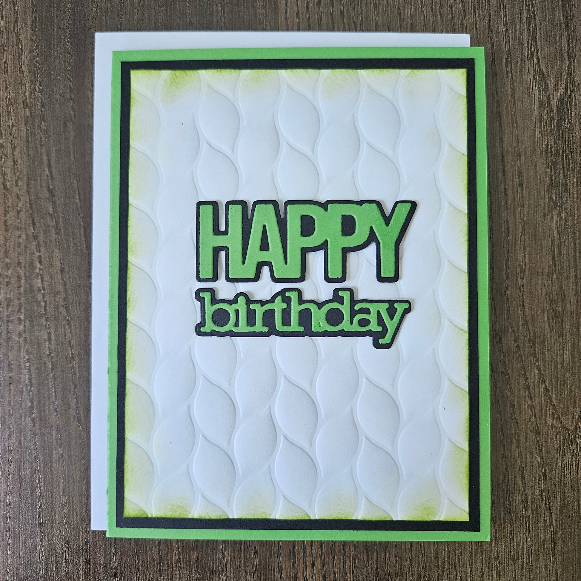 A handmade square greeting card with a white embossed wave pattern, featuring the words 'HAPPY birthday' in green and black. The card is framed with a green and black border and is photographed flat on a dark wood background.