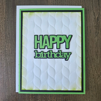 A handmade square greeting card with a white embossed wave pattern, featuring the words 'HAPPY birthday' in green and black. The card is framed with a green and black border and is photographed flat on a dark wood background.