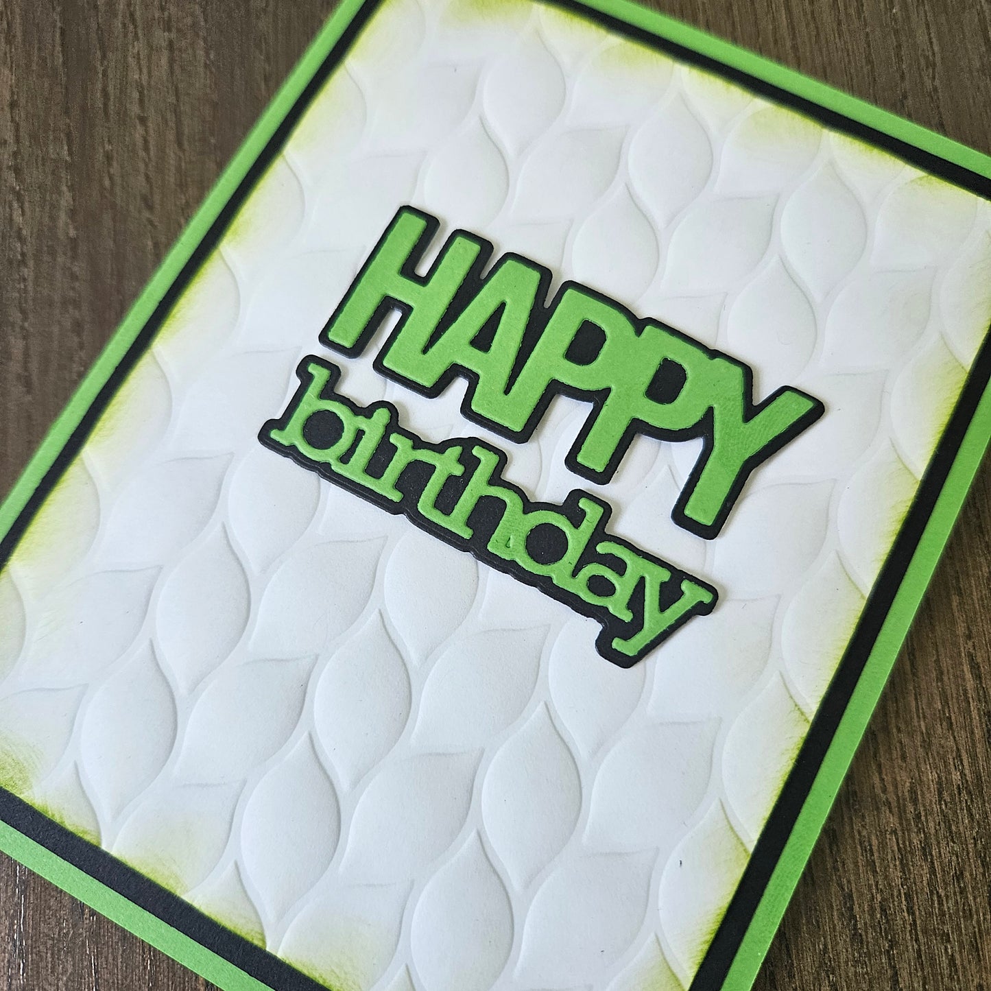 A handmade square birthday card with a white embossed wavy pattern, featuring 'HAPPY birthday' in green die-cut letters with black outlines, bordered in green and black, shown on a dark wood background.